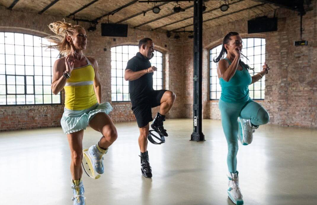 Three people exercising in a spacious room with large windows and brick walls.