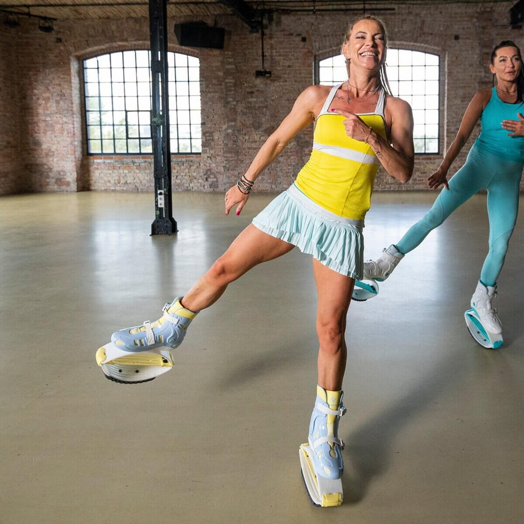 Two women exercising indoors while wearing blue and yellow Fit Boots Rebound Boots