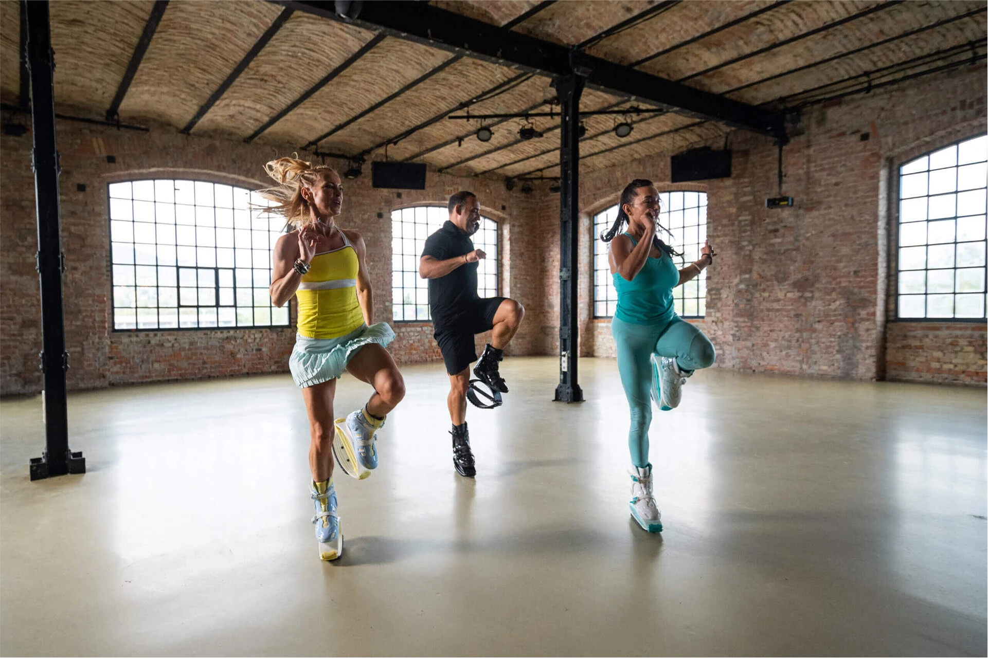 Three people exercising in a spacious room with brick walls and large windows.