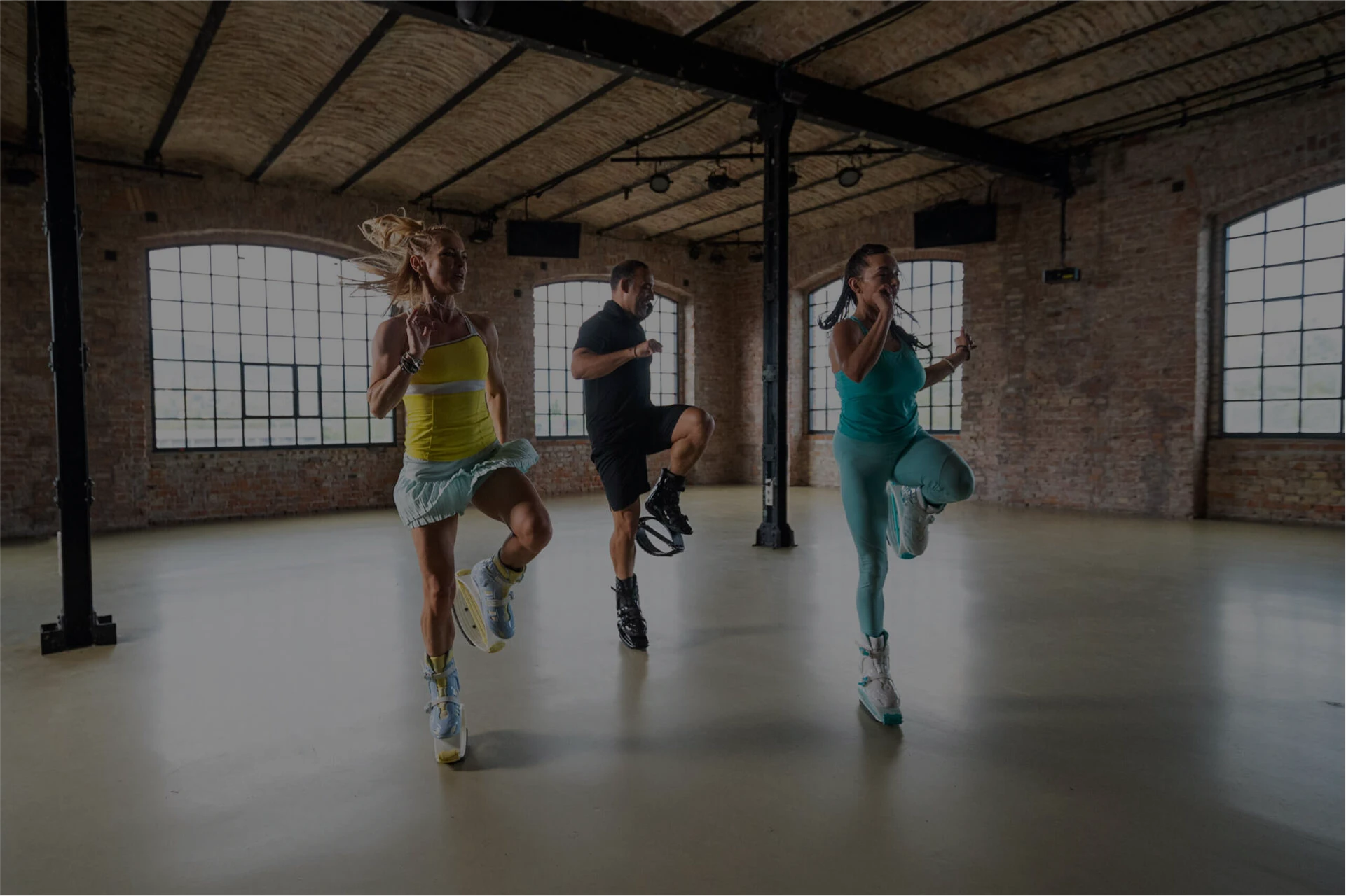 Three people exercising in a spacious room with brick walls and large windows.
