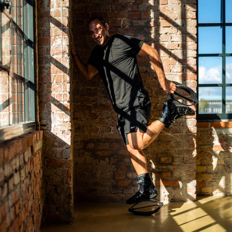 Homme adossé à un mur de briques, posant et s'étirant tout en portant des bottes de rebond Fit Boots à l'intérieur.