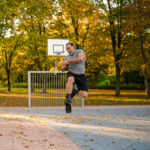 Homme sautant en l'air en portant des bottes de rebond Fit Boots sur un terrain de basket en plein air entouré d'arbres au feuillage d'automne.