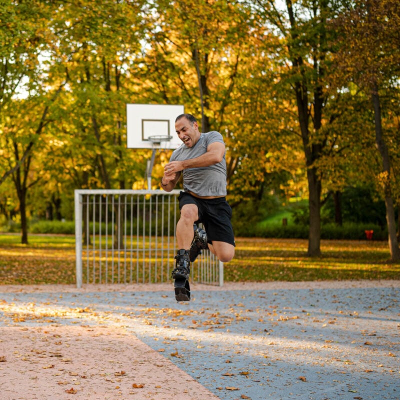 Hombre saltando en el aire mientras lleva puestas las botas Fit Boots Rebound Boots en una cancha de baloncesto al aire libre rodeada de árboles con follaje otoñal.